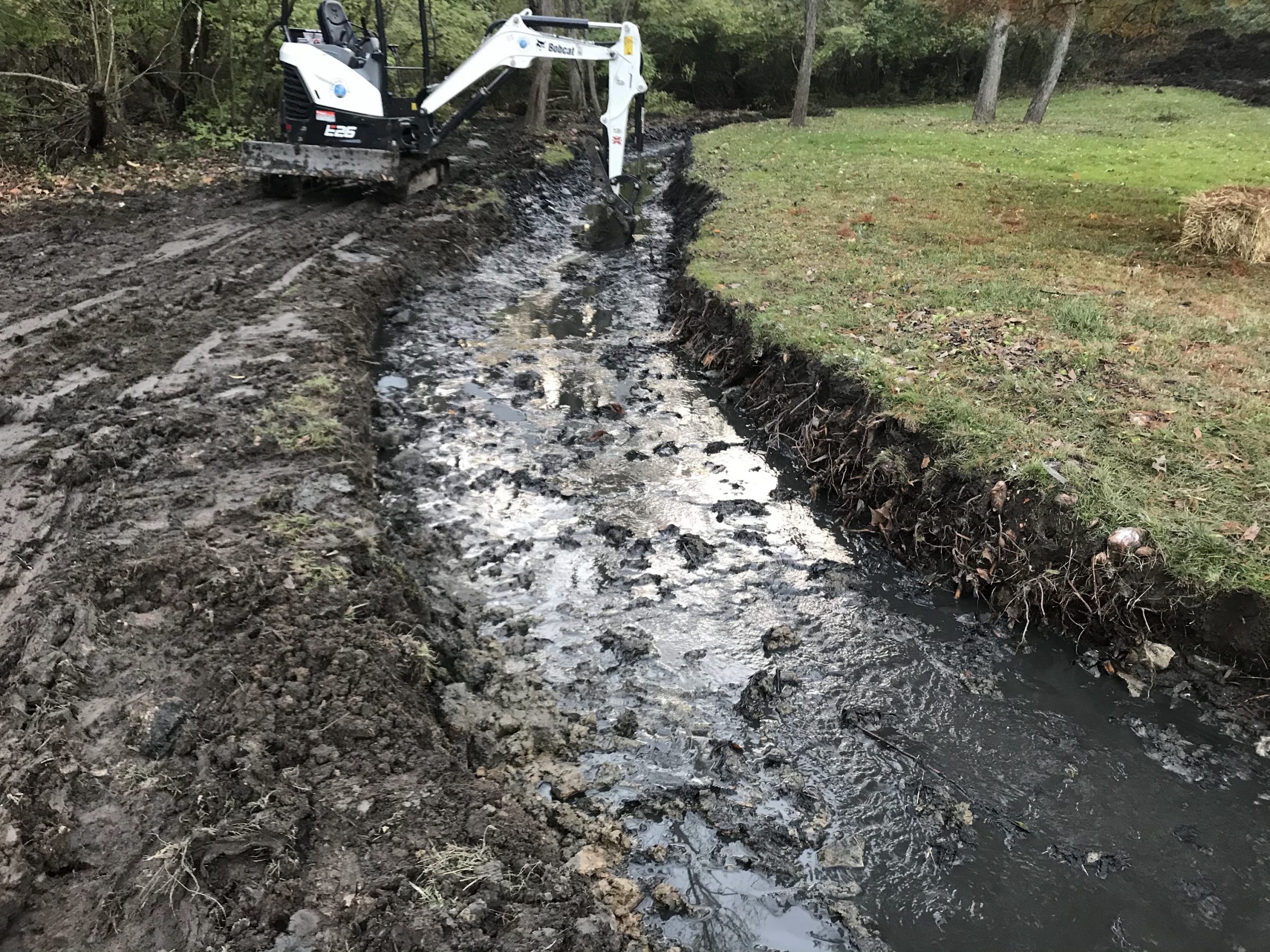 Excavator clearing muddy drainage ditch beside grassy field