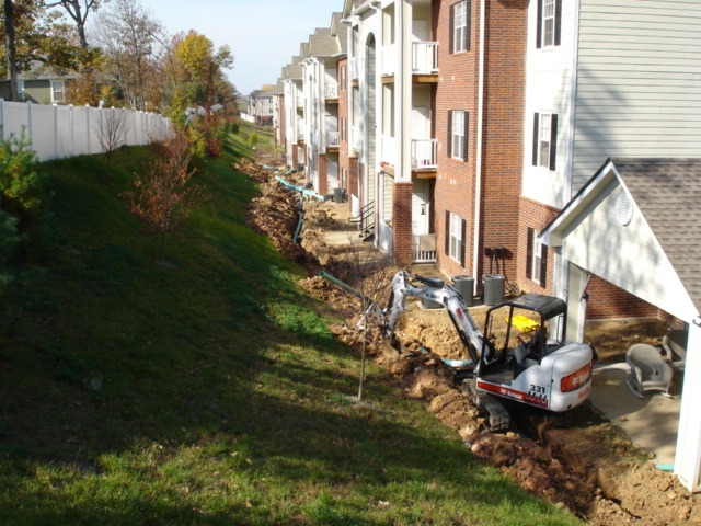 Excavator digging trench beside apartment buildings