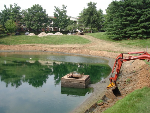 Excavator digging beside small pond near apartments