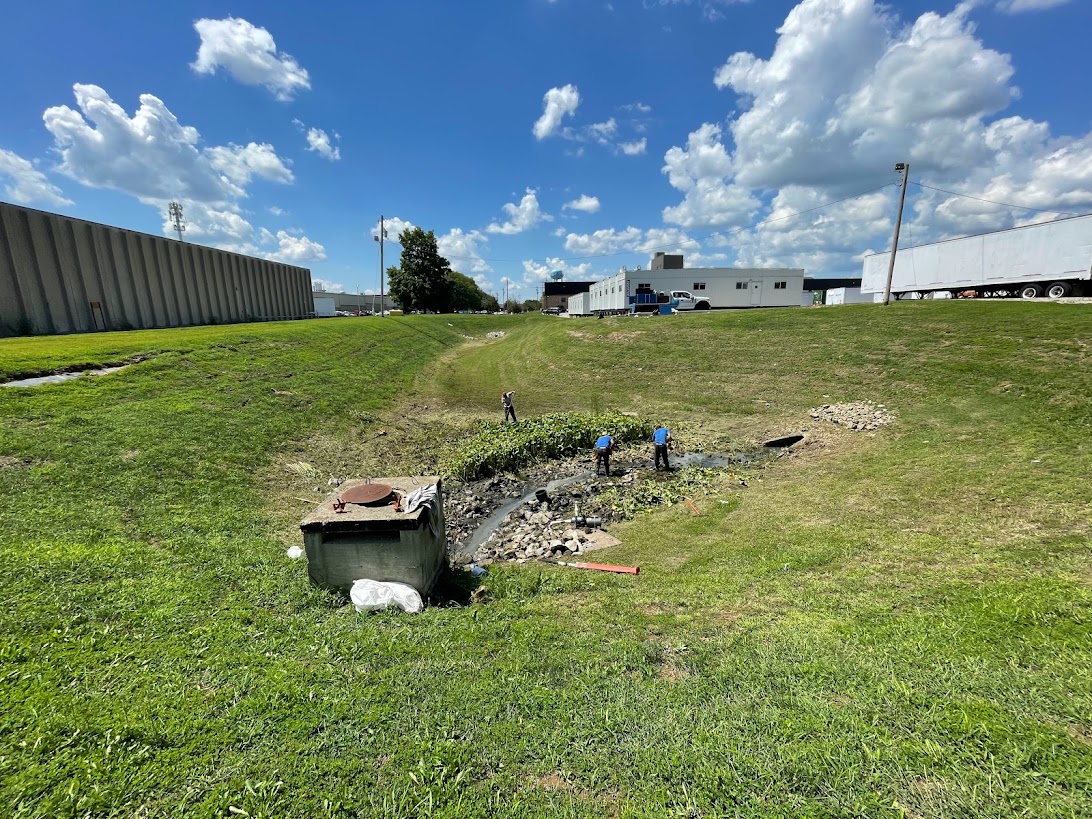 Workers clearing drainage ditch near industrial buildings